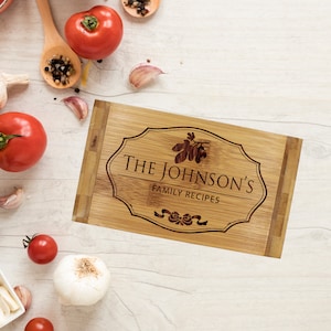 Pu&ograve; includere: Un tagliere rettangolare in bamb&ugrave; con un bordo decorativo inciso al laser, una piccola illustrazione di un rametto e il testo THE JOHNSON'S FAMILY RECIPES con un fiocco in basso, centrato sulla superficie in legno marrone chiaro.