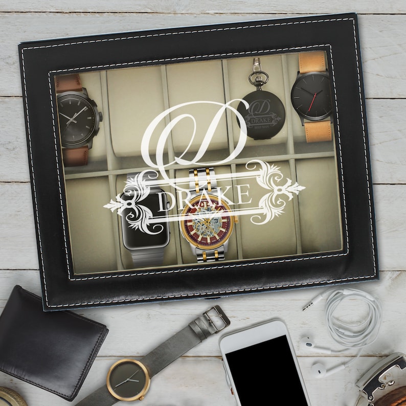 May include: Black leather watch display case with a clear top, holding several watches. The case is personalized with the name "Drake" and a large initial "D". Other accessories like a wallet, phone, and headphones are also visible.