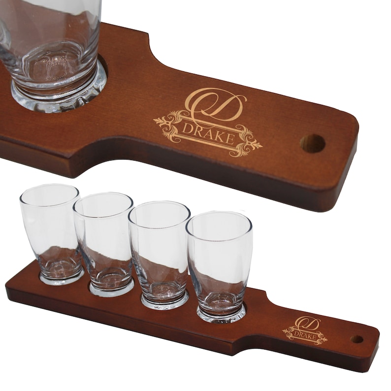 May include: Two wooden beer flight boards with clear glasses. One board holds four glasses, the other holds one. The boards are brown with the name "DRAKE" and a decorative design etched into the wood.