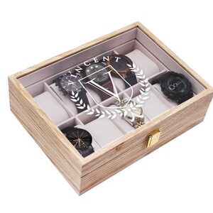 May include: A wooden watch box with a clear glass top, containing several watches. The box is light brown with a gold clasp. The watches have black, brown, and silver bands. The name "VINCENT" is printed on the glass.