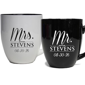 May include: Two ceramic mugs, one white and one black. The white mug has "Mrs. STEVENS 08-30-26" printed in black script. The black mug has "Mr. STEVENS 08-30-26" printed in white script. Both mugs have a curved shape and a handle.