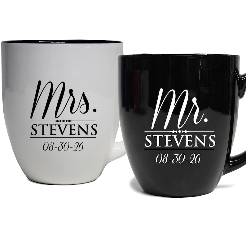 May include: Two ceramic mugs, one white and one black. The white mug has "Mrs. STEVENS 08-30-26" printed in black script. The black mug has "Mr. STEVENS 08-30-26" printed in white script. Both mugs have a curved shape and a handle.