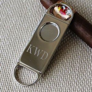 May include: Silver cigar cutter with a round blade and a ring for cutting. The cutter has the initials "KWD" engraved on the side.