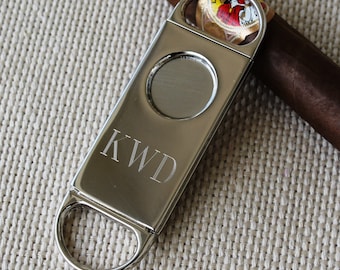 May include: Silver cigar cutter with a round blade and a ring for cutting. The cutter has the initials "KWD" engraved on the side.