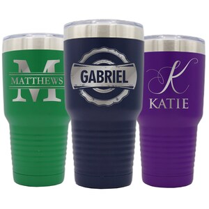 May include: Three stainless steel tumblers with personalized engravings. The tumbler on the left is green with the letter "M" and the name "MATTHEWS". The middle tumbler is blue with the name "GABRIEL" in a vintage style logo. The tumbler on the right is purple with the letter "K" and the name "KATIE".