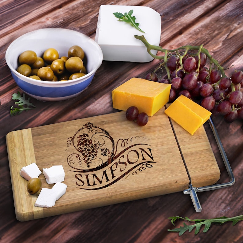 May include: Personalized bamboo cheese board with a wire cheese cutter. The board features a grapevine design and the name "SIMPSON" engraved on it. The board is surrounded by cheese, olives, grapes, and greenery.