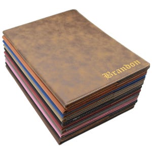 May include: A stack of personalized brown leatherette portfolios with the name "Brandon" embossed in gold lettering. The portfolios are in various colors, including brown, blue, pink, and black, with white stitching around the edges.
