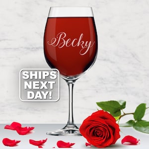 Puede incluir: Una copa de vino de cristal transparente llena de líquido rojo intenso, con el nombre Becky grabado en letra cursiva blanca en el cáliz. Una rosa roja vibrante con hojas verdes y pétalos de rosa dispersos se encuentra en la base de la copa.