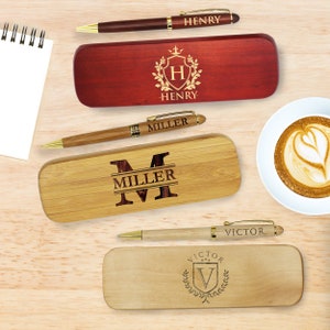 May include: Three personalized pen and box sets. The first set has a red box with a gold crest and the name "HENRY". The second set has a natural wood box with a gold initial "M" and the name "MILLER". The third set has a natural wood box with a gold crest and the name "VICTOR".