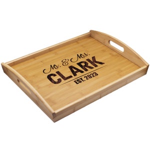 Puede incluir: Una bandeja de servicio rectangular de bamb&uacute; con bordes elevados y asas laterales integradas. El centro de la bandeja presenta un texto grabado a l&aacute;ser en color oscuro que dice Mr. & Mrs. CLARK EST. 2023 sobre la veta de madera clara.