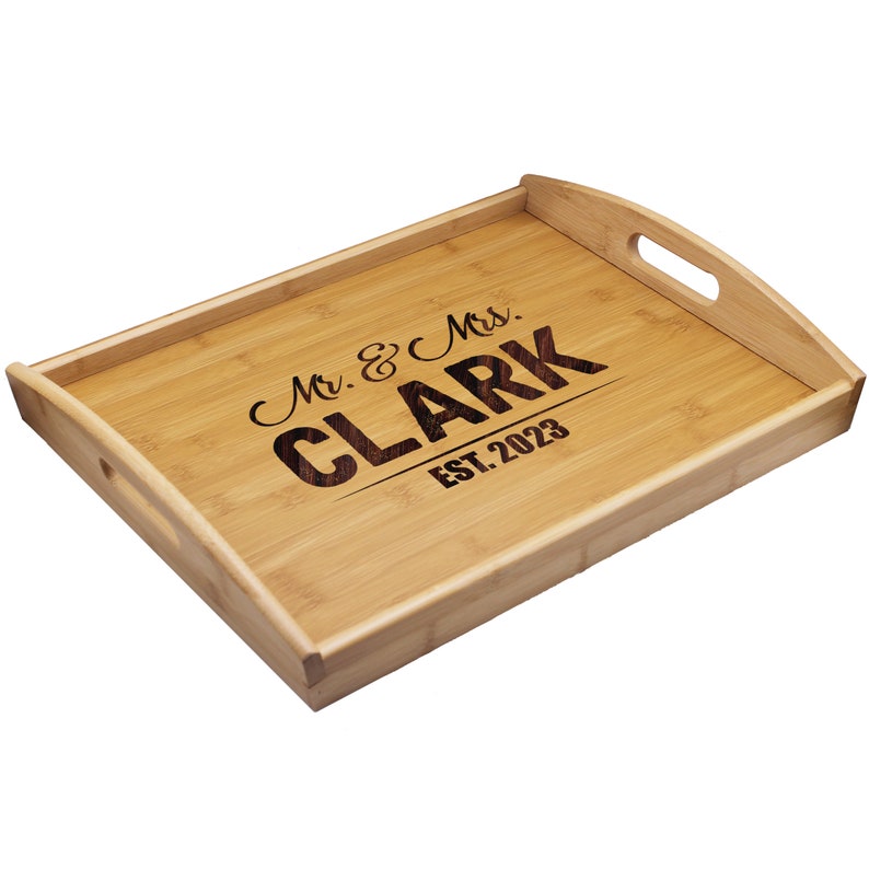Puede incluir: Una bandeja de servicio rectangular de bamb&uacute; con bordes elevados y asas laterales integradas. El centro de la bandeja presenta un texto grabado a l&aacute;ser en color oscuro que dice Mr. & Mrs. CLARK EST. 2023 sobre la veta de madera clara.