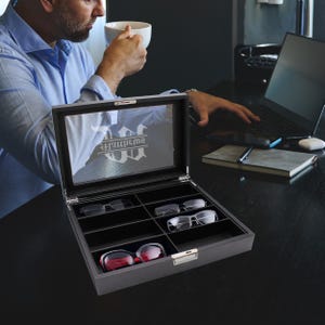 May include: A rectangular black storage case for eyewear with a clear glass lid featuring the white printed text Matthews. The interior is divided into eight individual compartments, with three pairs of glasses visible inside the organizer box.