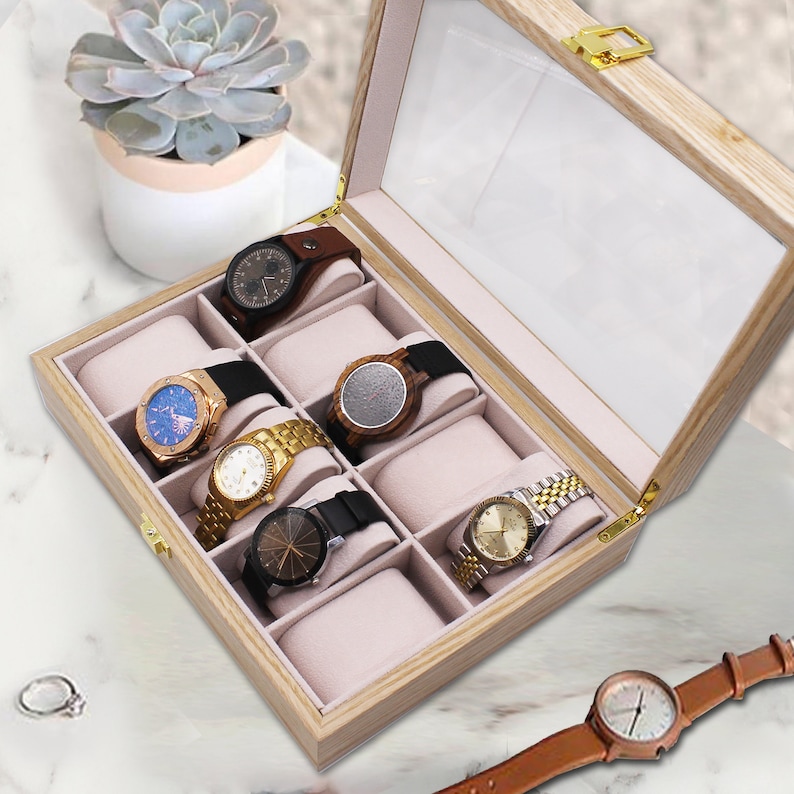 May include: A light-colored wooden watch display box with a clear glass lid and gold-tone metal hinges. The interior features eight individual compartments lined with soft beige fabric, each containing a watch cushion. Six various wristwatches are displayed.