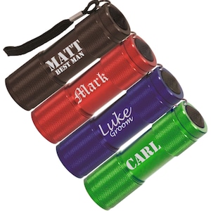 May include: Four cylindrical metal flashlights in black, red, blue, and green. Each flashlight features a textured grip and a black wrist strap. The black flashlight is engraved with MATT BEST MAN, the red with Mark, the blue with Luke Groom, and the green with CARL.