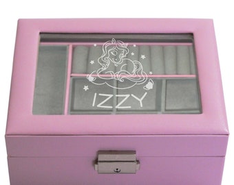 May include: A rectangular light pink jewelry box with a clear glass lid. The lid features a white graphic of a unicorn on a cloud surrounded by stars and the name IZZY printed below. The interior is lined with grey fabric and has compartments.