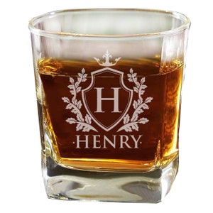 Pode incluir: Um copo de u&iacute;sque de vidro transparente com um design personalizado. O design apresenta um escudo com uma coroa no topo, a letra "H" dentro do escudo e folhas de carvalho que cercam o escudo. O nome "HENRY" est&aacute; gravado abaixo do escudo.