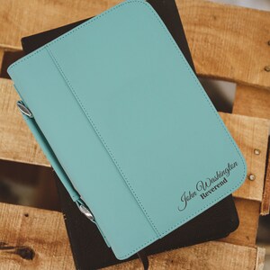 May include: A teal leather Bible cover with the name "John Washington, Reverend" printed in black script. The cover is closed and has a pen holder. The cover is resting on a black book, set against a wooden background.