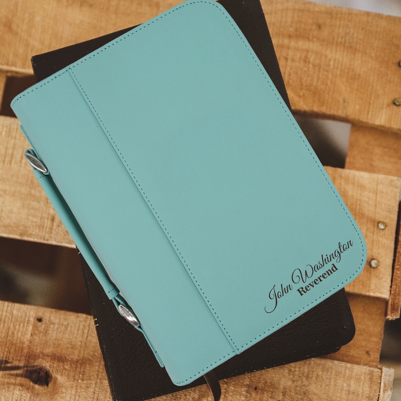 May include: A teal leather Bible cover with the name "John Washington, Reverend" printed in black script. The cover is closed and has a pen holder. The cover is resting on a black book, set against a wooden background.