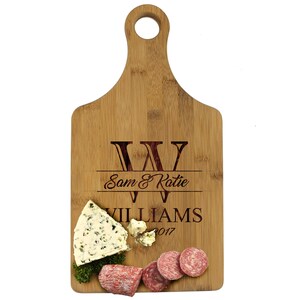 May include: A personalized bamboo cutting board with a handle. The board is engraved with the initials "WW", the names "Sam & Katie", the last name "Williams", and the year "2017". The board has a piece of blue cheese and slices of salami on it.