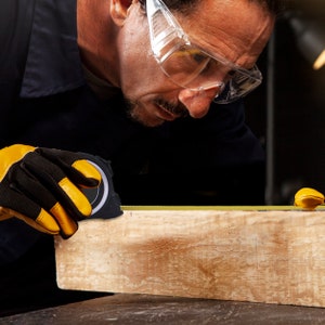 May include: A carpenter using a black and silver tape measure to measure a wooden plank. The carpenter wears yellow and black work gloves and clear safety glasses. The tape measure extends across the wood, showing the measurement.