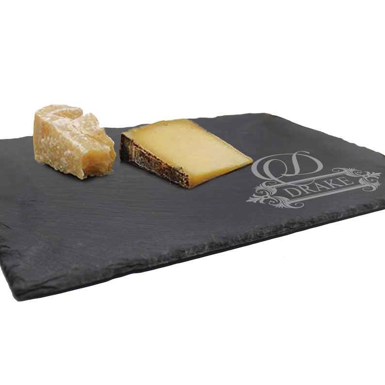 May include: A rectangular black slate serving board with rough, uneven edges. The surface features an engraved design with the letter D and the text DRAKE. Two pieces of yellow and beige cheese are placed on the left side of the dark stone board.