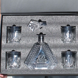 May include: A clear glass decanter with a diamond-cut pattern and a stopper, accompanied by four matching crystal glasses, all nestled in a black foam-lined box. The decanter features the engraved letter D and the name DRAKE on its front surface.