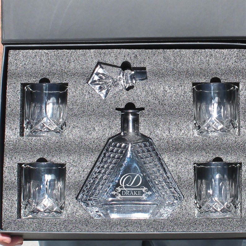 May include: A clear glass decanter with a diamond-cut pattern and a stopper, accompanied by four matching crystal glasses, all nestled in a black foam-lined box. The decanter features the engraved letter D and the name DRAKE on its front surface.