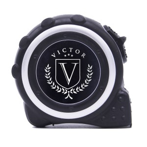 May include: Black tape measure with a silver and black face. The face features the word "VICTOR" above a shield with a "V" inside, surrounded by a laurel wreath. The tape measure is a construction tool.
