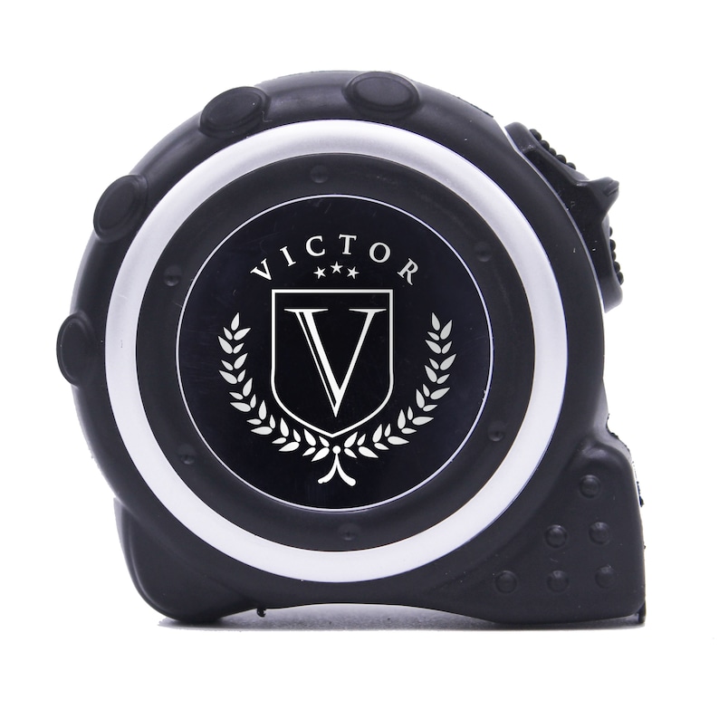 May include: Black tape measure with a silver and black face. The face features the word "VICTOR" above a shield with a "V" inside, surrounded by a laurel wreath. The tape measure is a construction tool.