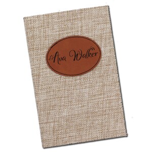 May include: A rectangular notebook with a textured, beige fabric cover. A brown, oval leather patch is affixed to the front, displaying the name "Ava Walker" in elegant script. The notebook's design suggests a personalized or custom item.
