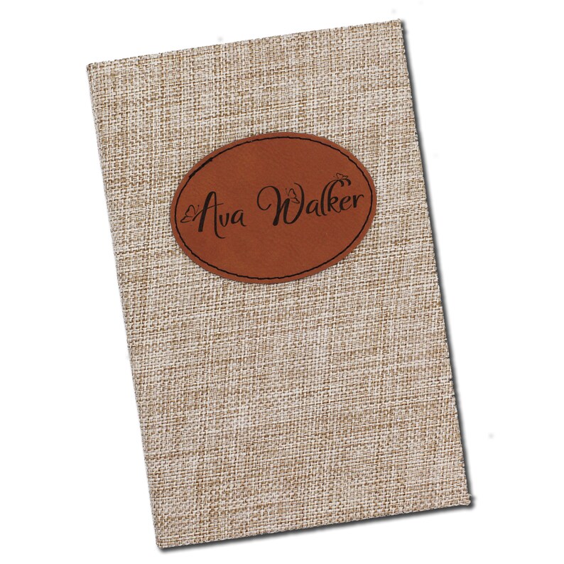 May include: A rectangular notebook with a textured, beige fabric cover. A brown, oval leather patch is affixed to the front, displaying the name "Ava Walker" in elegant script. The notebook's design suggests a personalized or custom item.