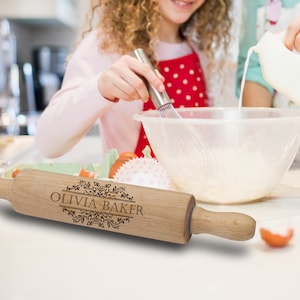 Puede incluir: Un rodillo de madera clara con la inscripci&oacute;n "OLIVIA BAKER" grabada en un marco decorativo. El rodillo est&aacute; delante de una persona que bate ingredientes en un bol. Se ve un delantal rojo con lunares blancos.