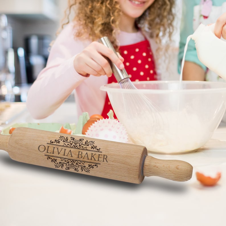 Puede incluir: Un rodillo de madera clara con la inscripci&oacute;n "OLIVIA BAKER" grabada en un marco decorativo. El rodillo est&aacute; delante de una persona que bate ingredientes en un bol. Se ve un delantal rojo con lunares blancos.