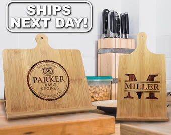 May include: Two light-colored bamboo wood recipe stands with laser-engraved designs. The left stand features a circular logo with a pretzel icon and the text Parker Family Recipes. The right stand displays a large letter M with the name Miller.