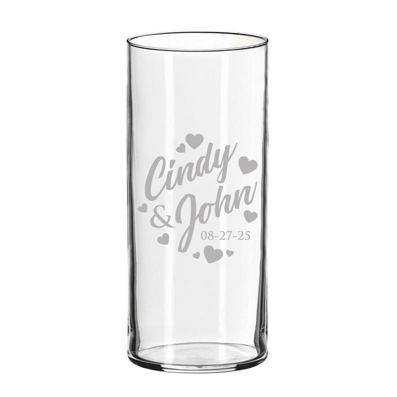 Op de afbeelding: Een cilindervormige vaas van helder glas met een gegraveerd ontwerp met de namen Cindy & John, de datum 08-27-25 en verschillende kleine hartvormen rondom de tekst. Het glas heeft een glad, transparant oppervlak en een rechte, verticale vorm.