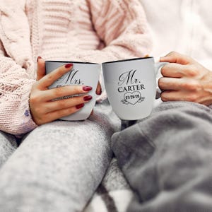 May include: Two white ceramic mugs with black rims. One mug reads "Mrs." and the other reads "Mr. Carter" with a heart and date. The mugs are held by two people, suggesting a couple. The mugs are a wedding or anniversary gift.