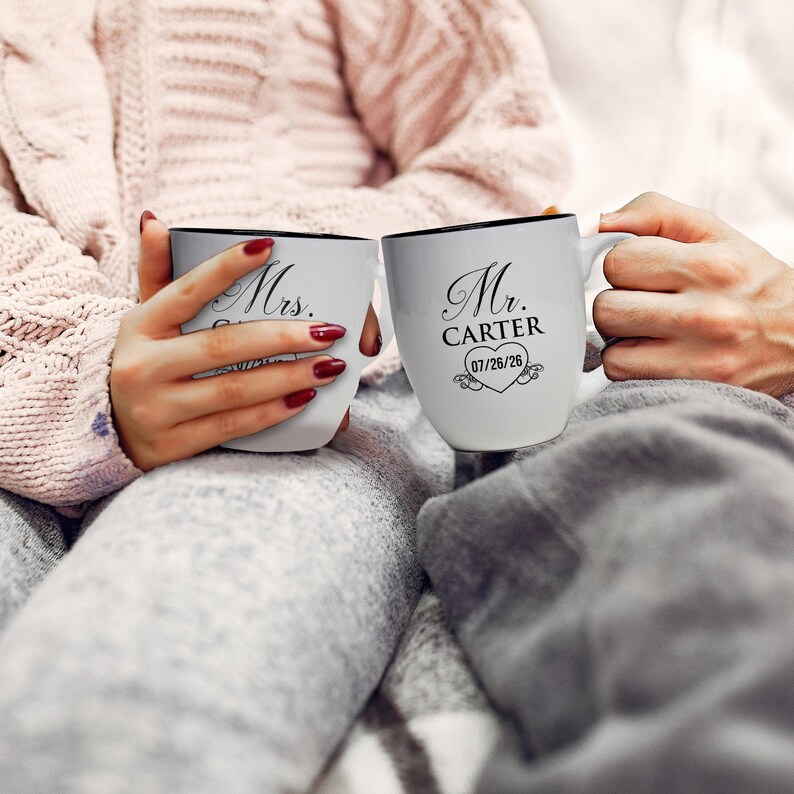 May include: Two white ceramic mugs with black rims. One mug reads "Mrs." and the other reads "Mr. Carter" with a heart and date. The mugs are held by two people, suggesting a couple. The mugs are a wedding or anniversary gift.