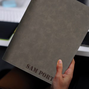 May include: A rectangular, dark gray portfolio folder with a smooth, matte finish and visible stitching along the edges. The name SAM PORTY is embossed in a dark, bold, sans-serif font near the bottom right corner of the front cover.