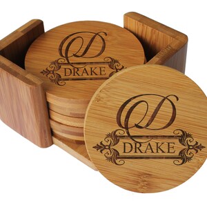 May include: A set of round bamboo coasters stored in a matching bamboo holder. Each coaster features a laser-engraved design with a large script letter D above a decorative frame containing the text DRAKE in a clean, capitalized serif font.
