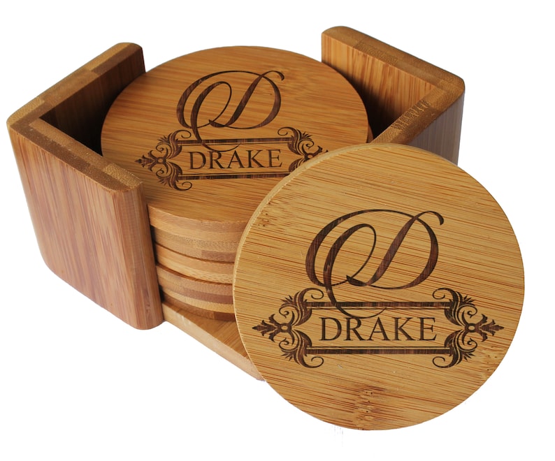 May include: A set of round bamboo coasters stored in a matching bamboo holder. Each coaster features a laser-engraved design with a large script letter D above a decorative frame containing the text DRAKE in a clean, capitalized serif font.
