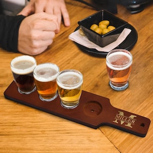 May include: A wooden beer flight paddle with four glasses of beer in varying shades of amber and brown. The paddle has a handle with the name "MILLER" engraved on it. A small black bowl of snacks sits nearby.