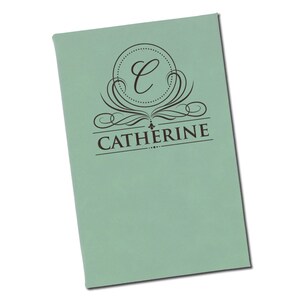Puede incluir: Un cuaderno verde claro personalizado con el nombre "Catherine" impreso en marr&oacute;n oscuro. El dise&ntilde;o incluye un monograma circular con la letra "C" y adornos decorativos.