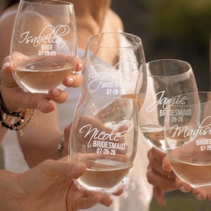 May include: Five clear stemless wine glasses filled with a light-coloured liquid. Each glass features white script text displaying a name, a wedding role such as Bride, Maid of Honour, or Bridesmaid, and the date 07-26-26 etched on the glass surface.