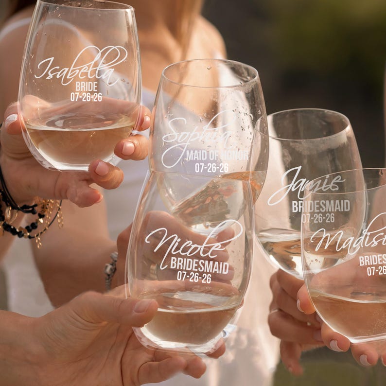 May include: Five clear stemless wine glasses filled with a light-coloured liquid. Each glass features white script text displaying a name, a wedding role such as Bride, Maid of Honour, or Bridesmaid, and the date 07-26-26 etched on the glass surface.