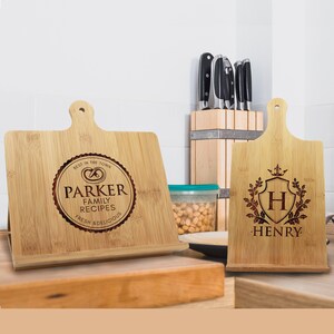 May include: Two light brown bamboo wood recipe stands with handles. The left stand features a circular engraving with a pretzel icon and the text "PARKER FAMILY RECIPES BEST IN THE TOWN FRESH & DELICIOUS". The right stand shows a shield with the letter "H" and "HENRY" below.