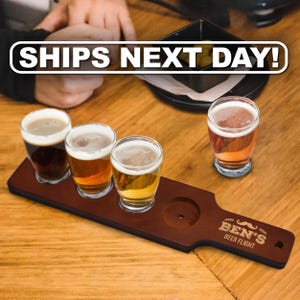 May include: A dark brown wooden beer flight paddle with a handle, featuring the engraved text BEN'S BEER FLIGHT and a mustache icon. It holds three clear glass beer samplers filled with different colored ales, with a fourth glass placed nearby.