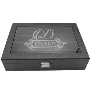 May include: A rectangular black storage box with a textured finish and a clear glass lid. The lid features an etched design with a large letter D and the name DRAKE centered within an ornate frame. A silver metal latch secures the front closure.