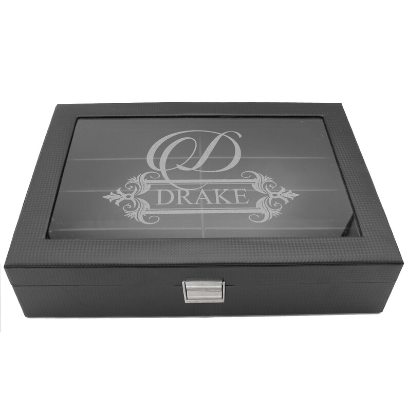 May include: A rectangular black storage box with a textured finish and a clear glass lid. The lid features an etched design with a large letter D and the name DRAKE centered within an ornate frame. A silver metal latch secures the front closure.