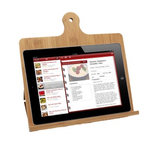 May include: A light brown bamboo tablet stand shaped like a cutting board with a handle and a hole at the top. A black tablet is resting on the stand, displaying a digital recipe for Raspberry Amaretto Tarts with a list of ingredients and instructions.