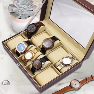 May include: A brown leather watch box with a glass top, filled with various wristwatches. The watches have different band and face designs, including gold, wood, and black leather. A succulent plant in a white pot is in the background.